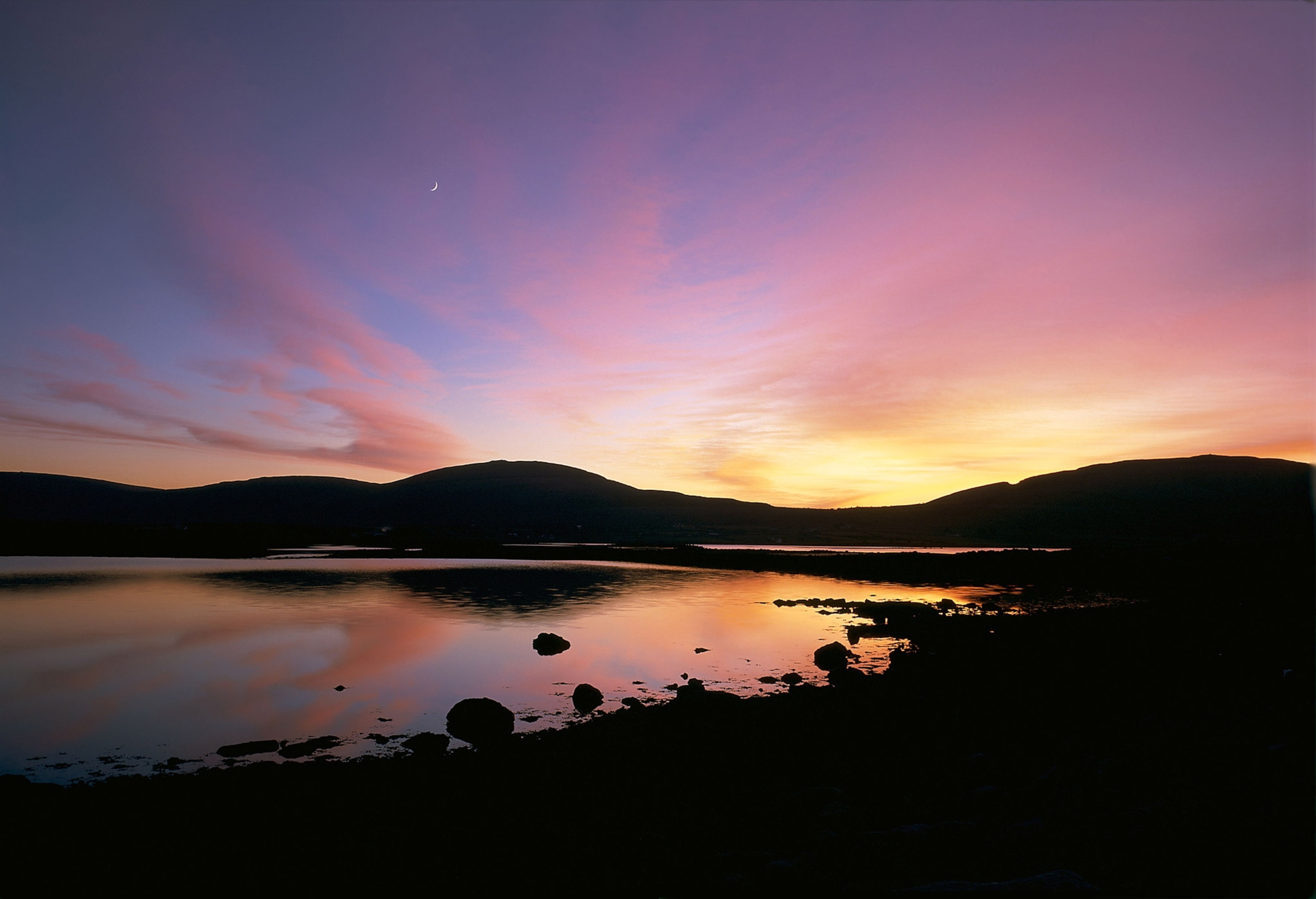 A pink sunset is reflected over a glassy lake surrounded by hills