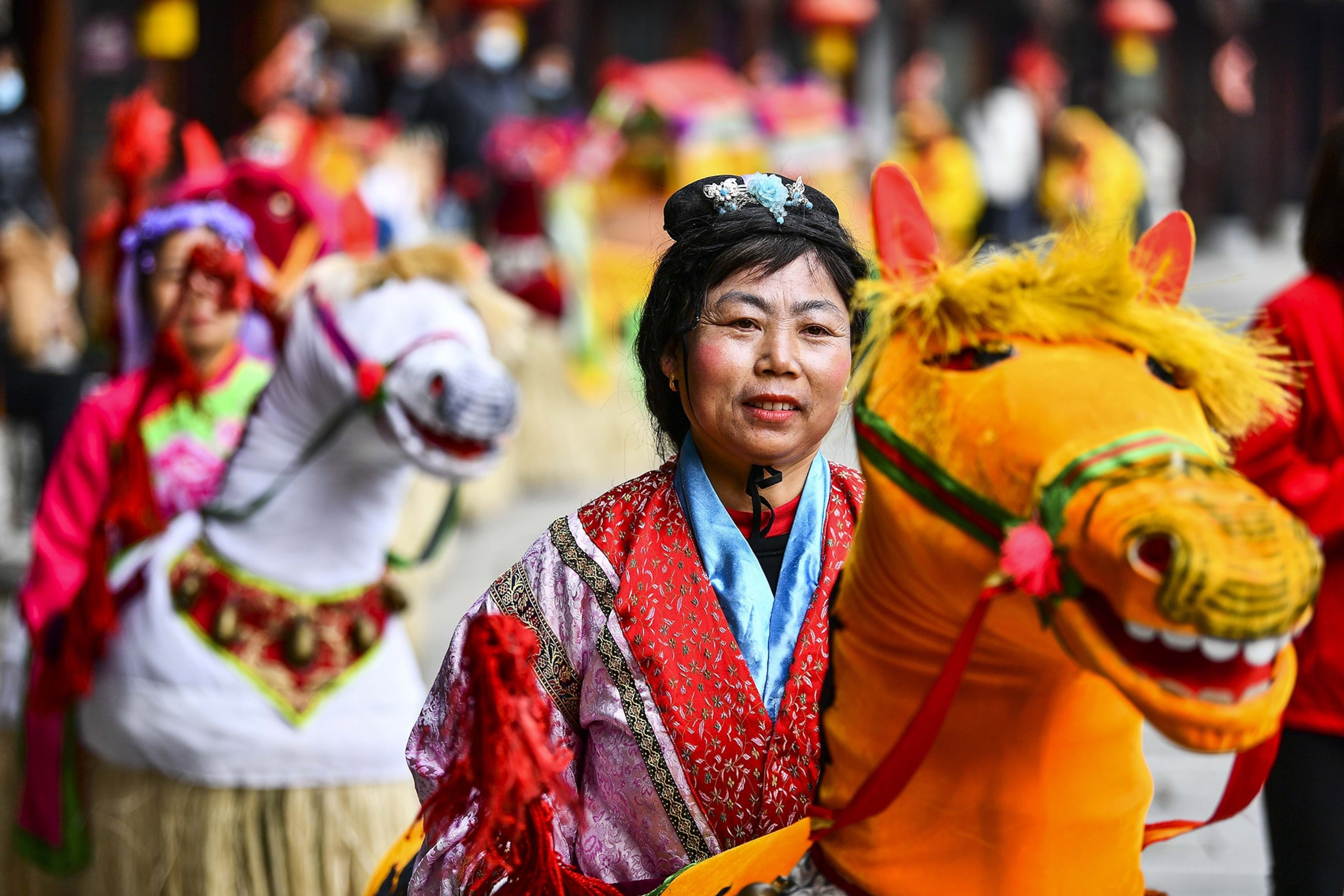 A woman in celebratory Chinese clothing astride a fake horse, surrounded by others on parade