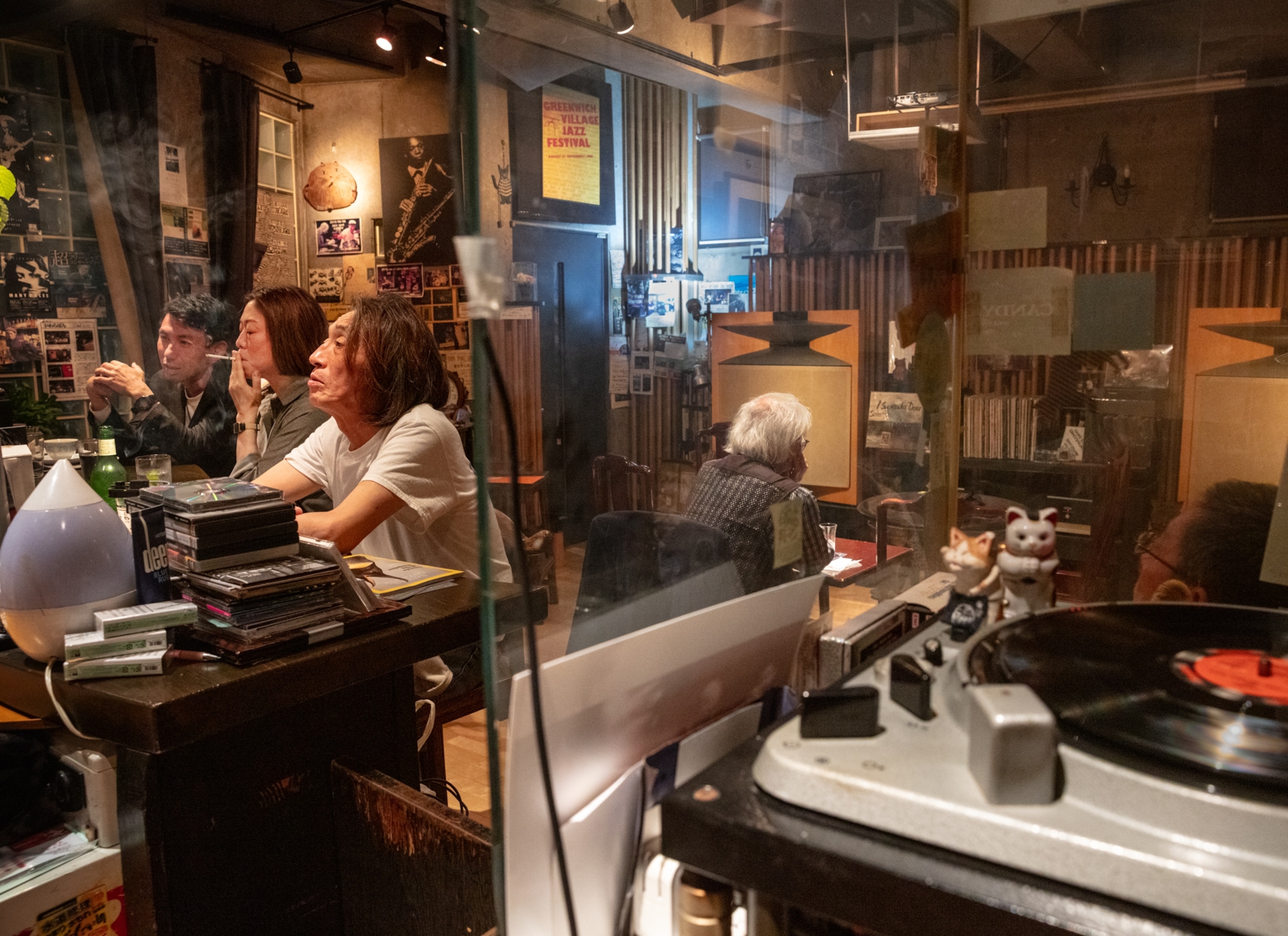 People smoking and drinking at the table loaded with books in the small room with record player on the foreground.