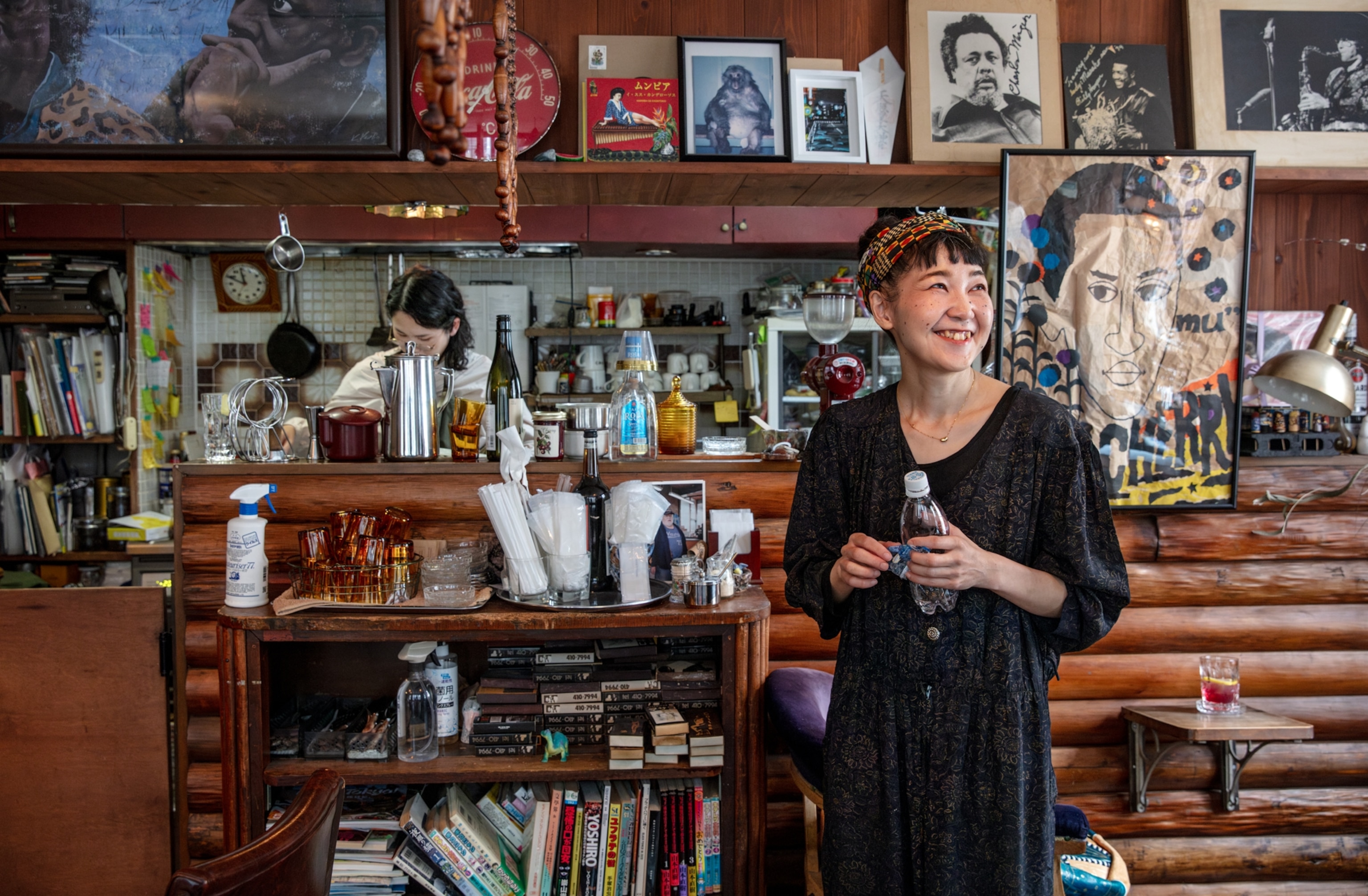 One women smiling and another one is working behind kitchen bar made of logs in the room decorated with photos, posters, books and records.