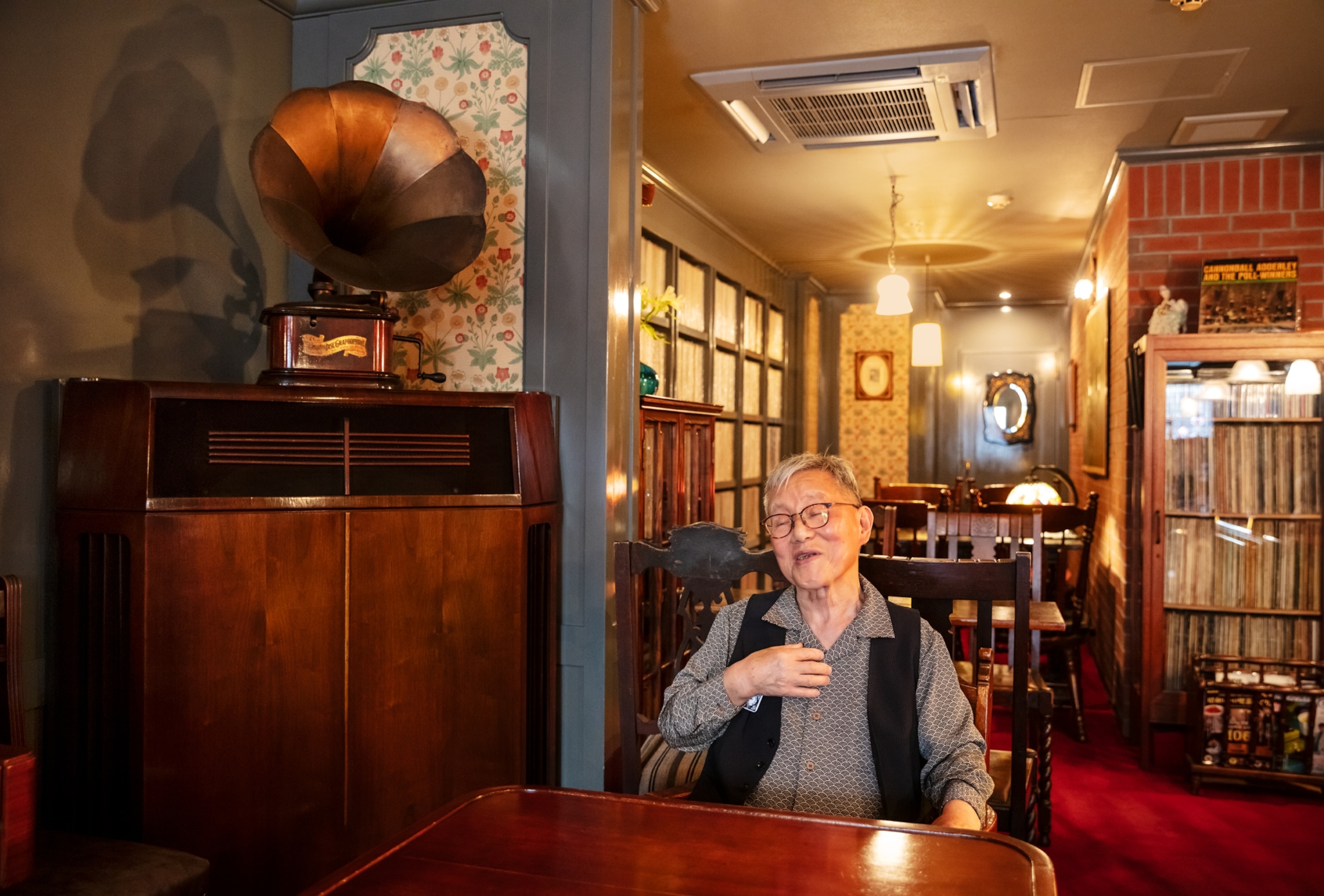 Man is glasses talking with his eyes closed and right hand on his chest. Gramophone displayed in the corner.