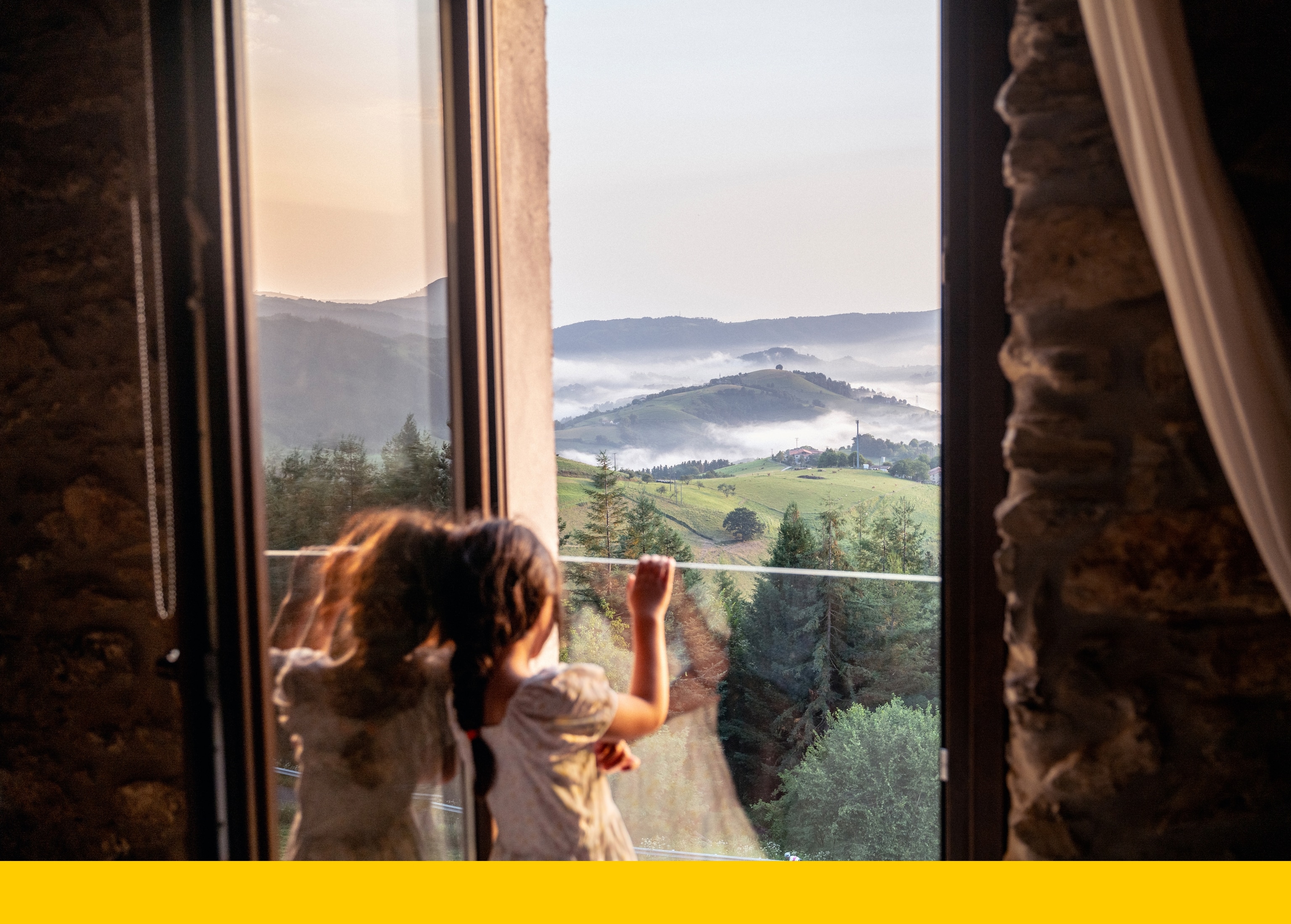 A young girl gazes out of a window toward the valleys and mountains surrounding the Basque village of Aia, where low clouds drift across the hills, softening the rugged landscape. 