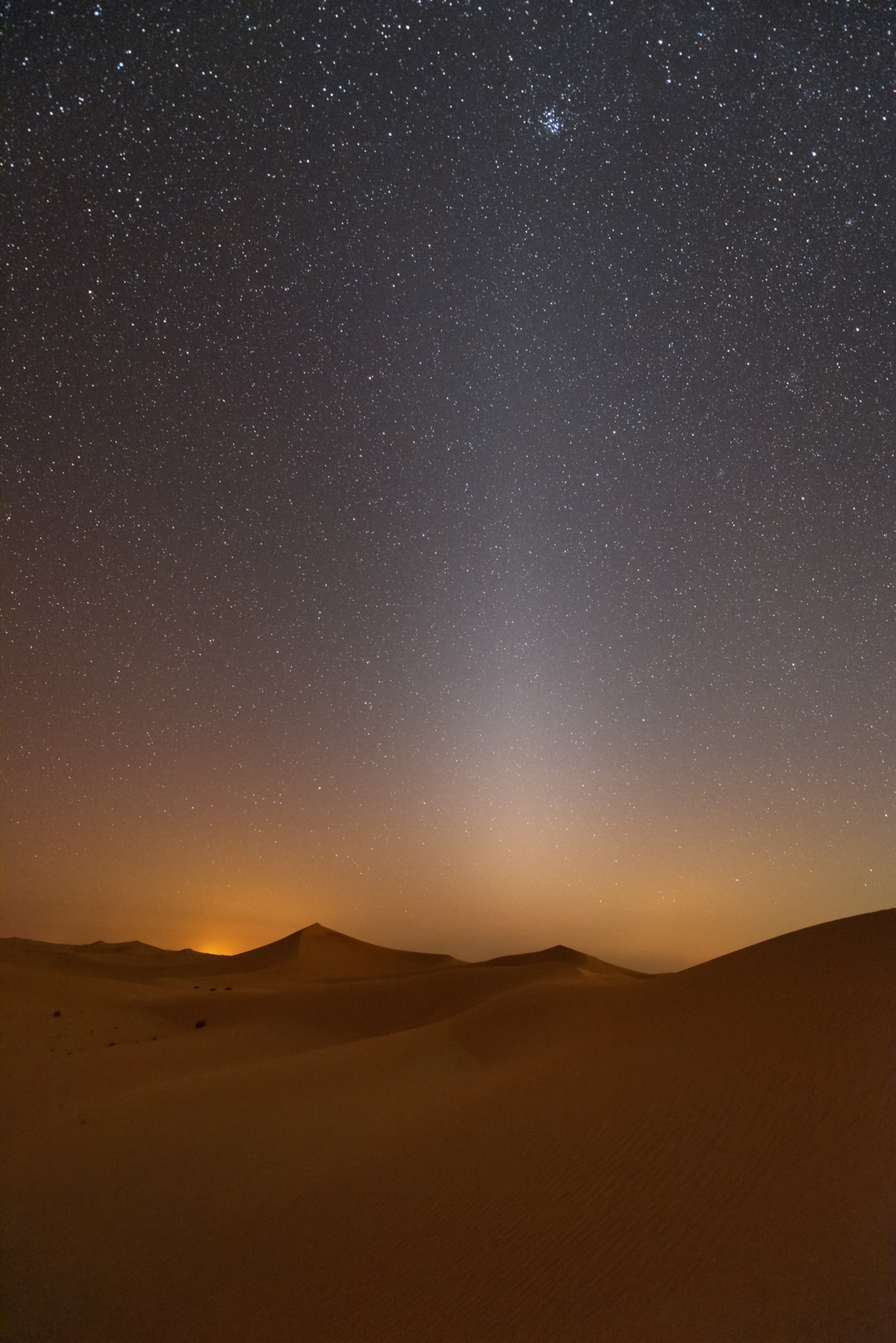 Starry desert nightscape with a glowing twilight horizon, sand dunes in foreground, and a faint vertical light beam rising into the star-filled sky.
